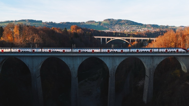 Double-deck train crossing a stone arch viaduct, autumn woods and a second bridge behind.