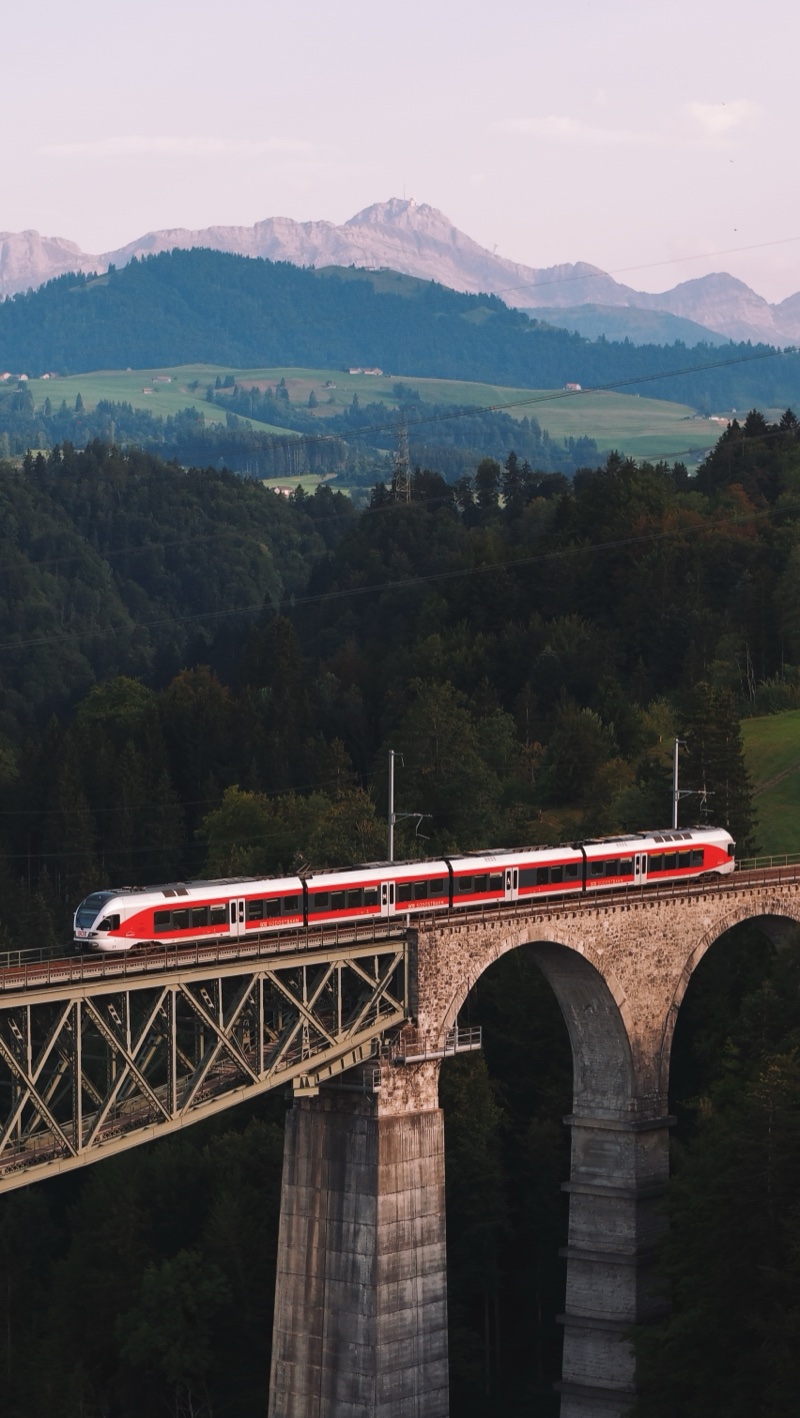 Red and white train crossing a viaduct of stone arches and a steel truss at dusk, mountains behind.