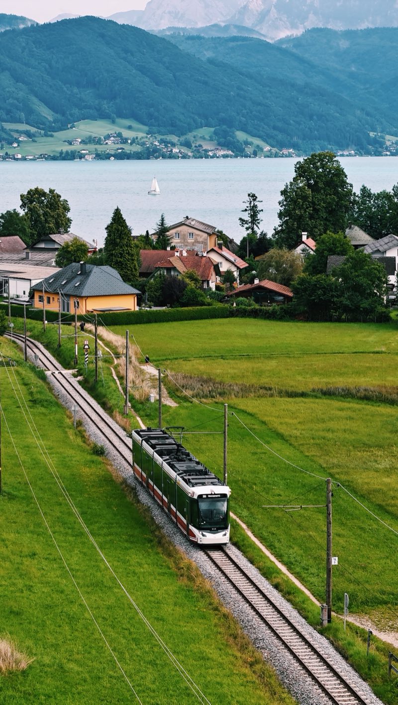 Green and white tram on a single track through meadows, with a lake and mountains behind.