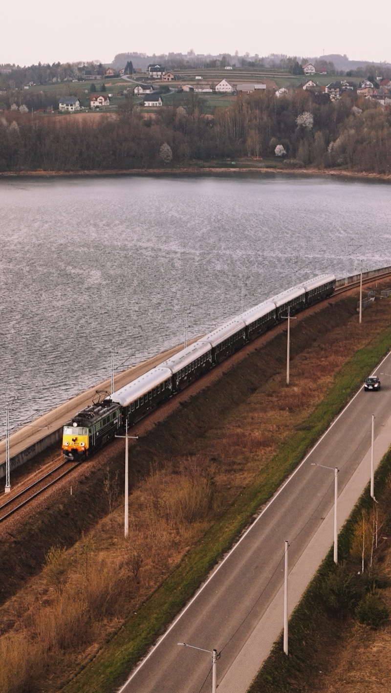 Passenger train on an embankment between a lake and a road, in evening light.