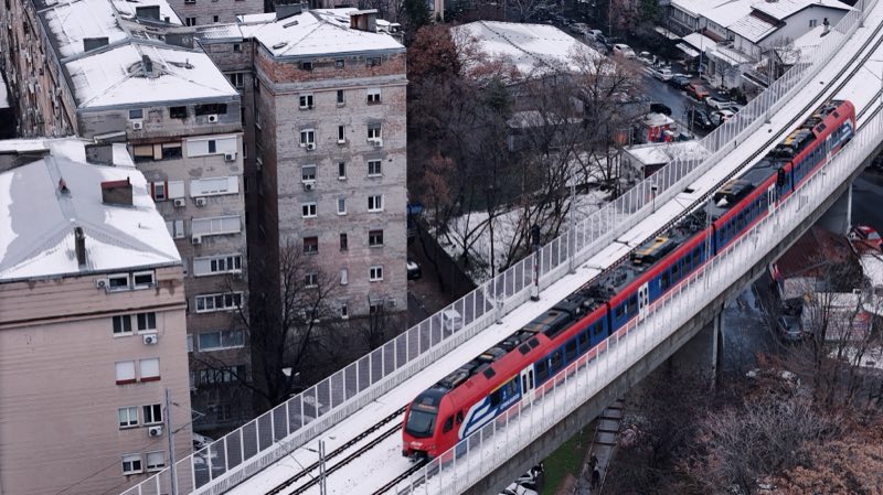 Red and blue train on an elevated line passing apartment blocks, snow on the roofs.