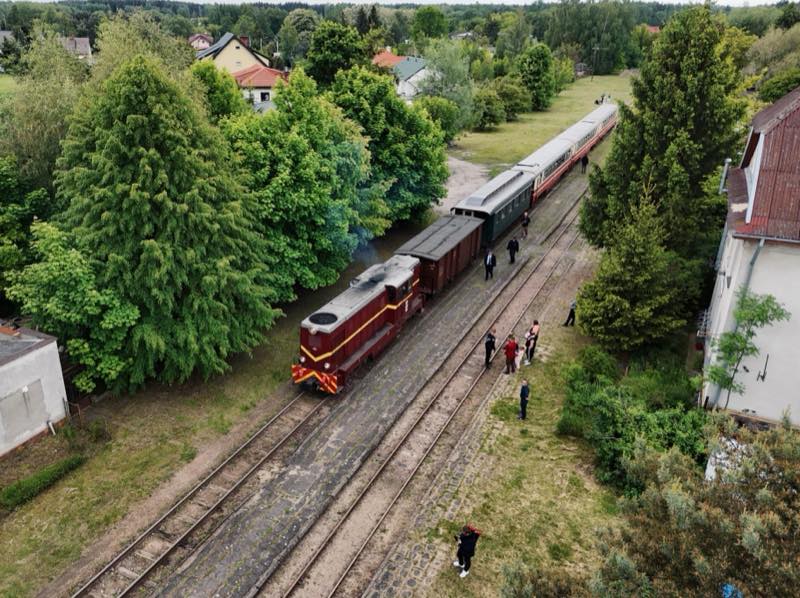 Red diesel locomotive hauling vintage carriages past trees, people watching beside the track.