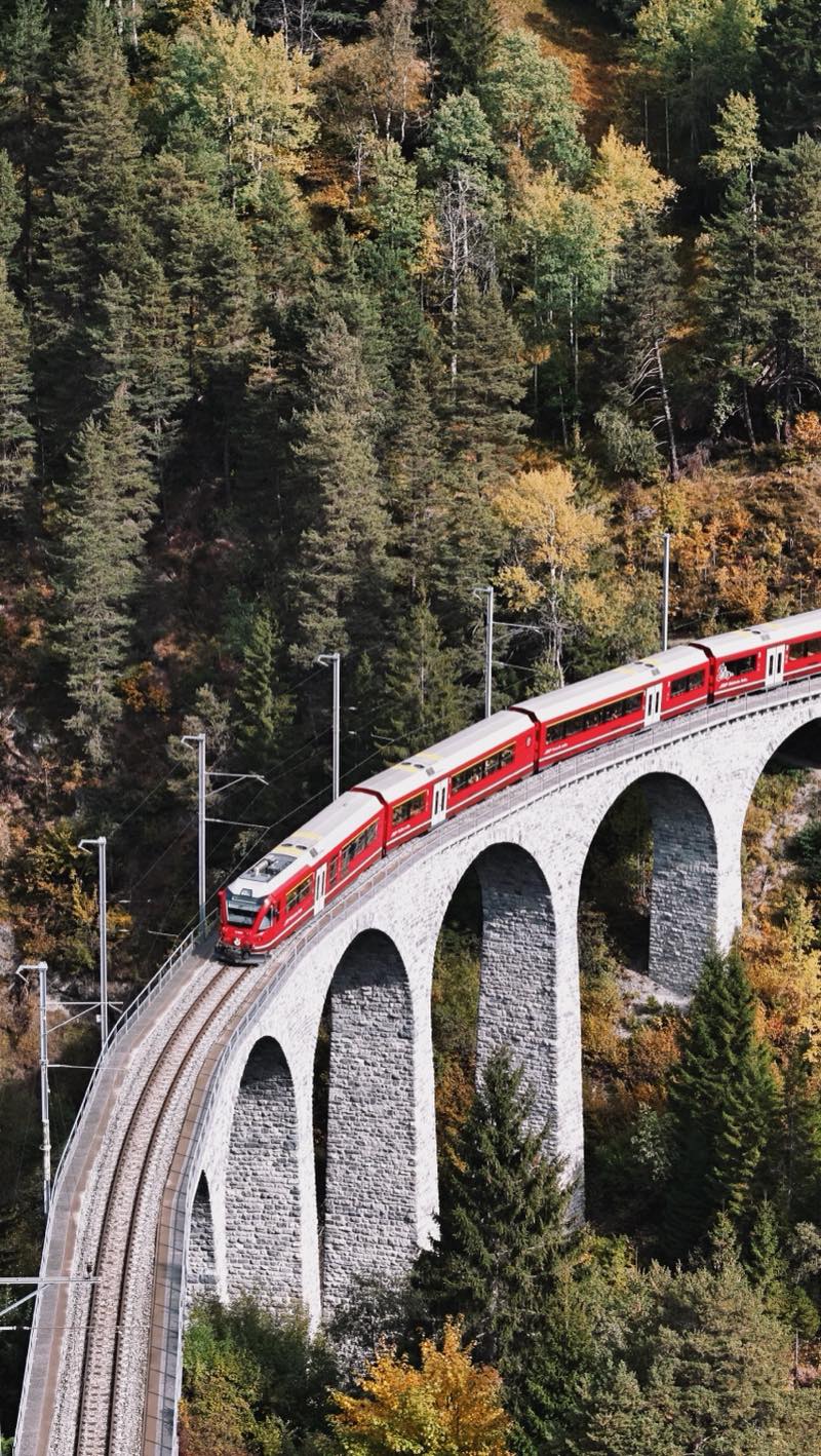 Red train on a curving stone viaduct against a forested slope.