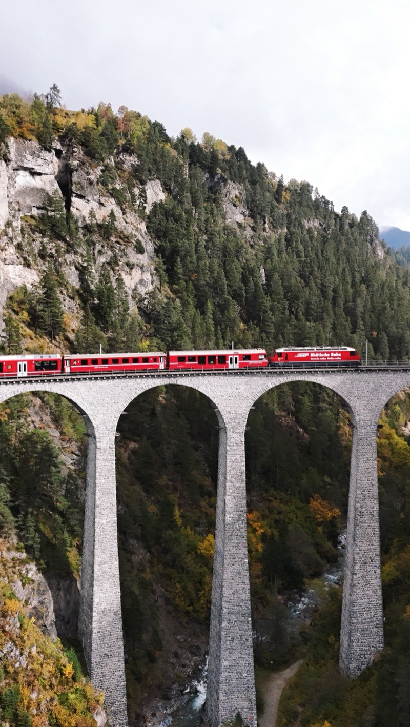 Red train on a tall stone viaduct, cliffs and forest behind.