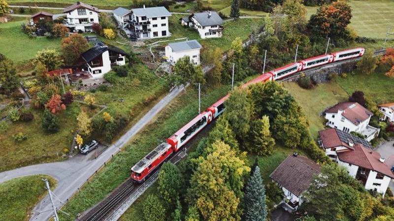 Red train winding through a village of chalets and autumn trees.
