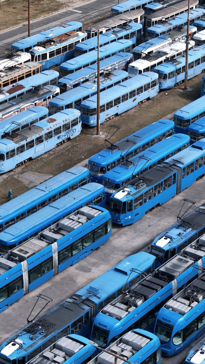 Rows of blue trams, old and new, parked closely in a depot yard, seen from above.