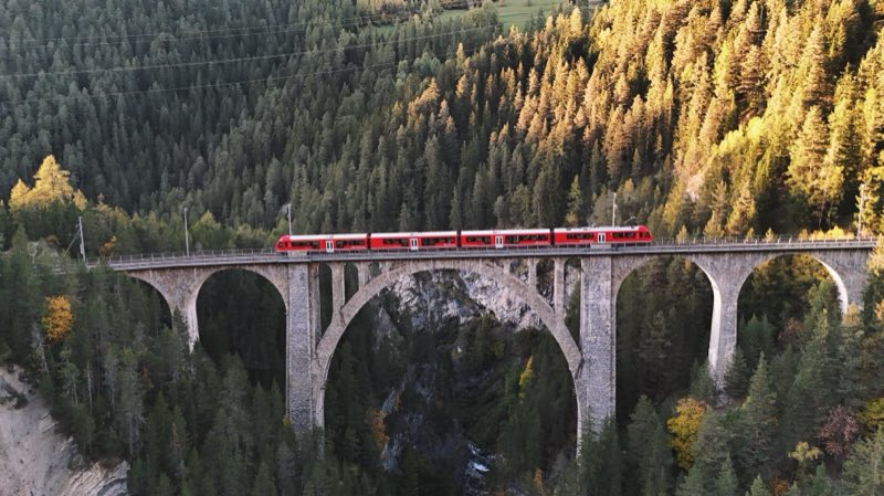 Short red train on a stone viaduct with one large central arch over a gorge, lit by low sun.
