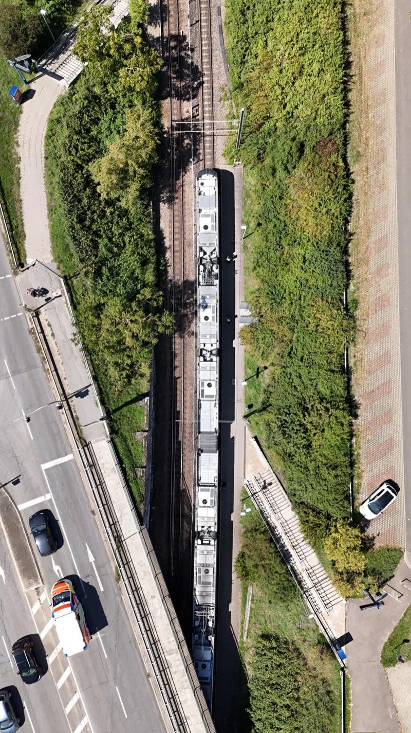 Train seen from directly above in a narrow corridor between a road and fields.