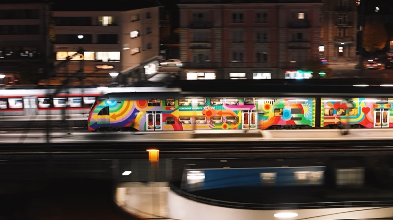 Train painted in bright graffiti colours passing a lit street at night.