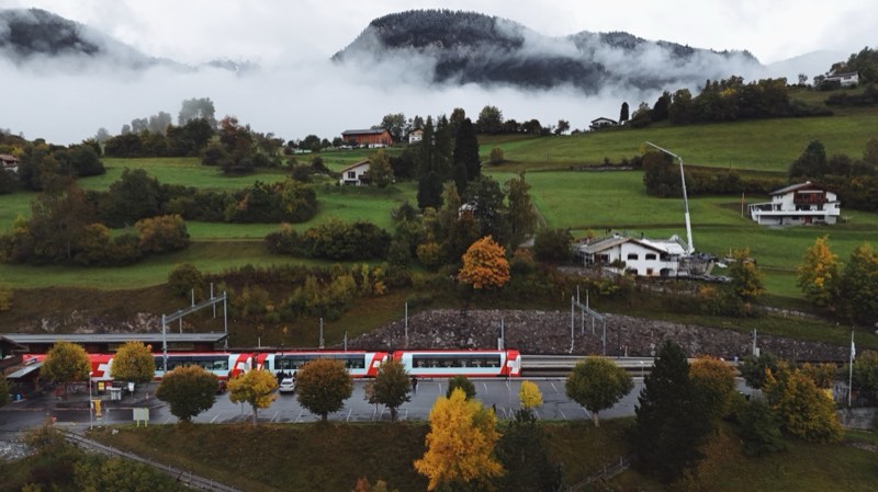 Train passing a row of autumn trees, low cloud on the mountains behind.