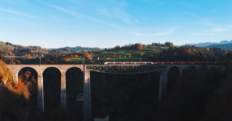 Two trains meeting on a long viaduct of stone arches and a steel truss, above a dark valley and green hills.